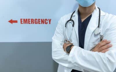 A doctor in a white coat and mask stands with arms crossed near a wall sign indicating the direction to the emergency room.