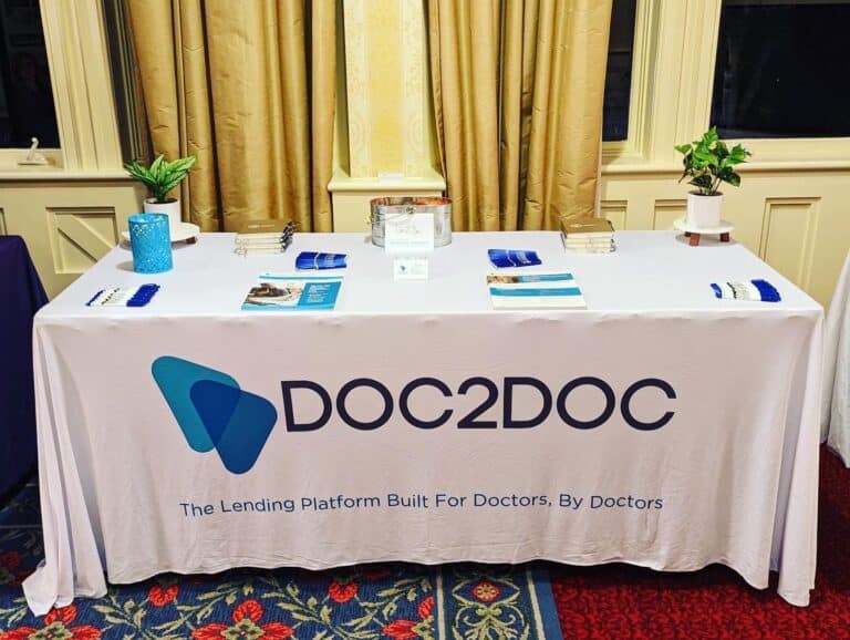 A booth with a table covered by a white tablecloth displaying "DOC2DOC," a lending platform for doctors, with brochures, business cards, and a donation box on top.