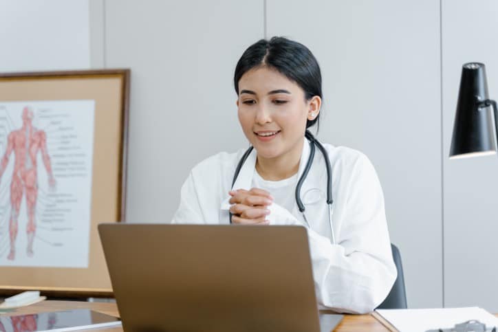 A doctor with a stethoscope around their neck using a laptop at a desk, with a framed anatomical diagram in the background.