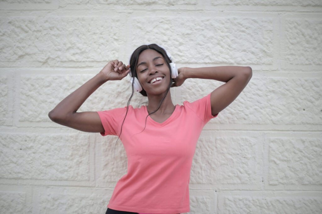A person wearing a pink shirt and headphones stands smiling against a light-colored brick wall, with eyes closed and arms raised.