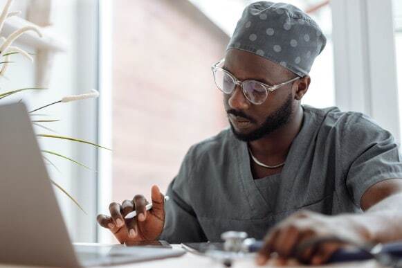 A person in medical scrubs and a polka dot cap is sitting at a desk, using a laptop and a digital pen.