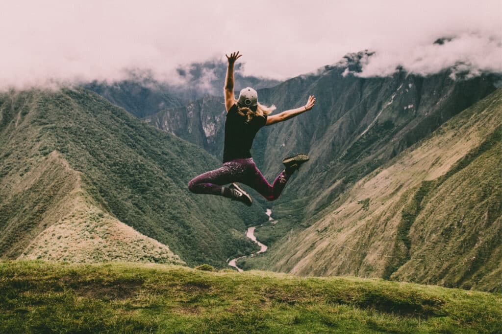 Person jumping in the air on a grassy hill with mountains and a river in the background, under a cloudy sky.