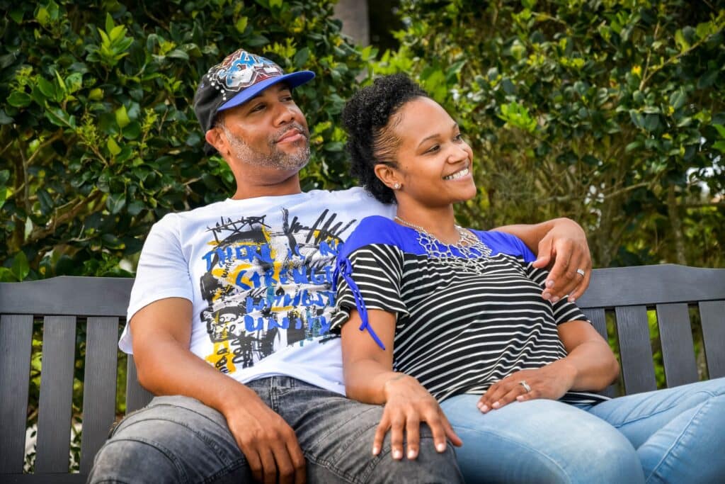 A couple sits on a bench outdoors, smiling and looking to the right.