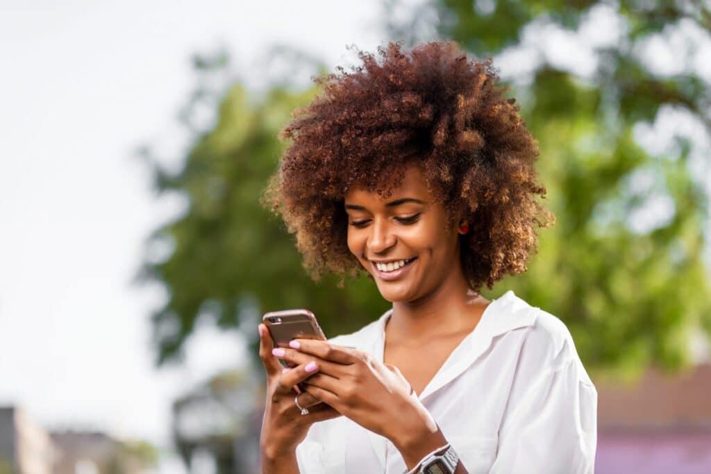 A woman with curly hair is smiling and looking at her phone while standing outside, with trees blurred in the background.