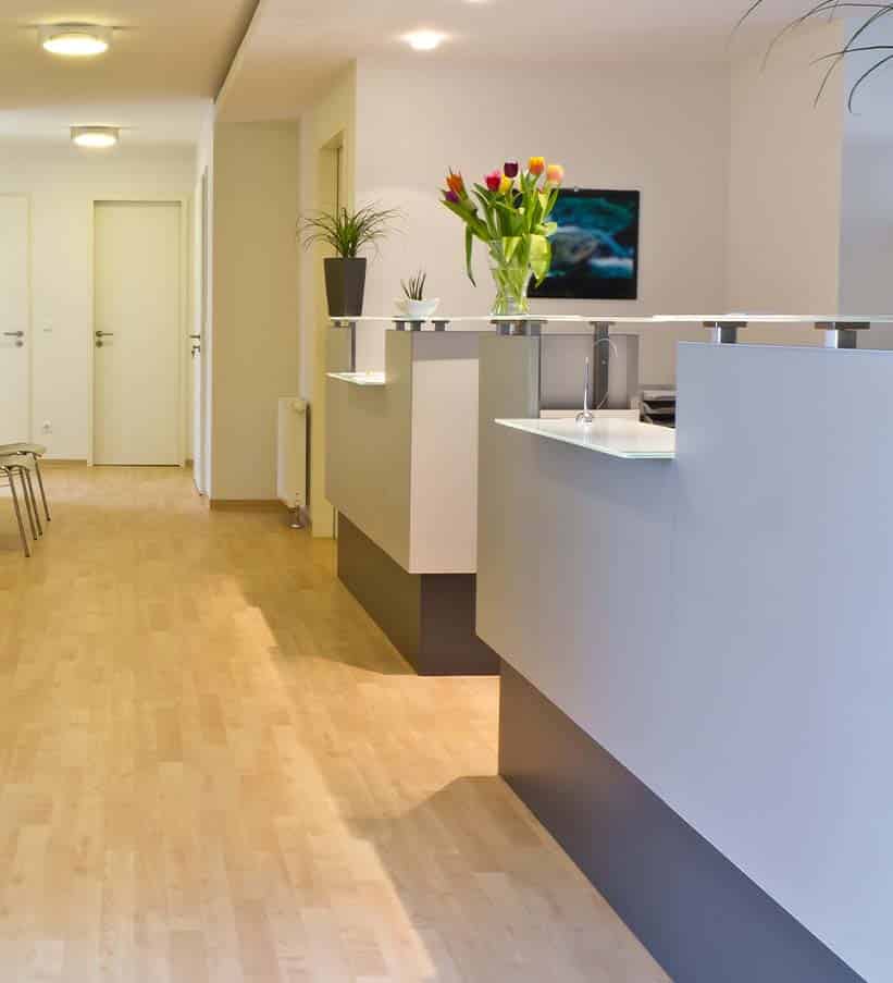 A modern, minimalist reception area with white counters, a vase of tulips, wooden floors, and closed doors along the corridor.