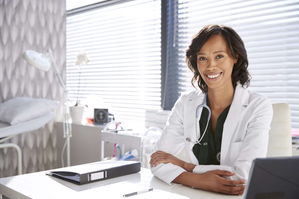 A young woman wearing a white lab coat and a stethoscope around her neck is sitting at a desk and smiling.