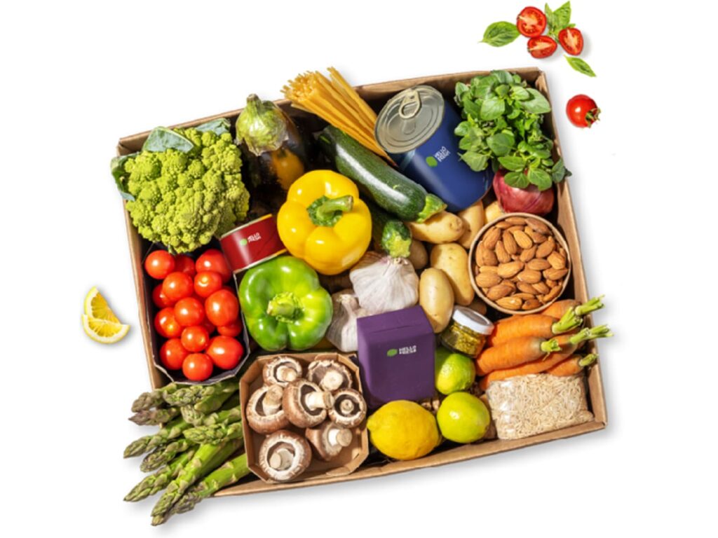 A neatly arranged box of assorted groceries including vegetables, canned goods, almonds, pasta, and herbs.