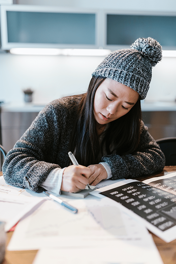 A person wearing a gray knit hat and sweater is writing at a table cluttered with papers