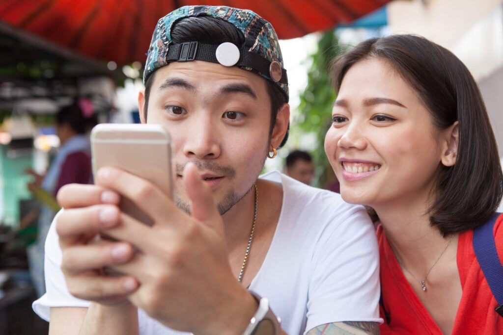 Two people sitting under an umbrella, looking at a smartphone.