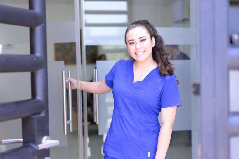 A woman in blue medical scrubs holds open a glass door, smiling at the camera.