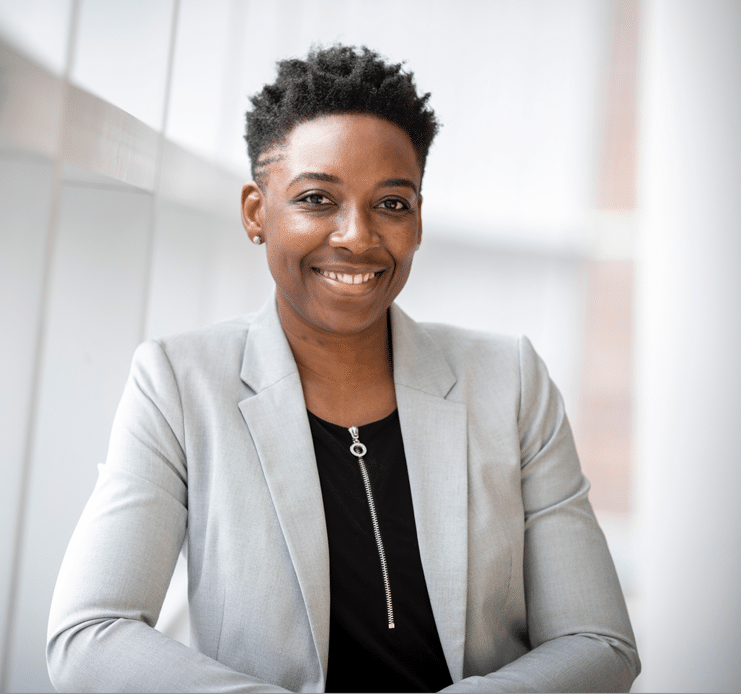 Person wearing a light gray blazer over a black top, smiling and standing against a bright, modern background.