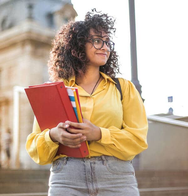 A person wearing glasses and a yellow shirt holds a red binder and books, standing outside near a building.