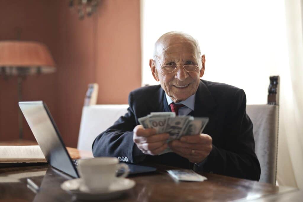 An elderly man wearing a suit sits at a table holding a stack of money, with an open laptop in front of him and a cup and saucer nearby.