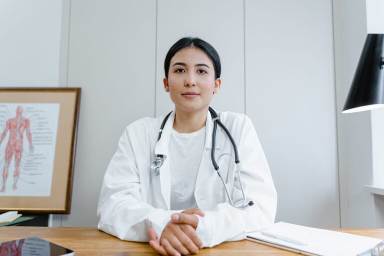 A doctor wearing a white coat and stethoscope sits at a desk, hands clasped, with medical charts beside her and a framed anatomical diagram on the wall in the background.