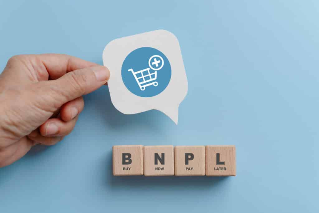 BNPL, buy now pay later, on wooden cube blocks and hand hold real white speech bubble paper cut with shopping cart icon on blue background