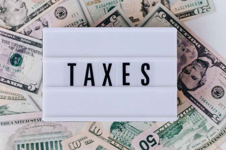 A white lightbox displaying the word "TAXES" is placed on a scattered array of various US dollar bills.