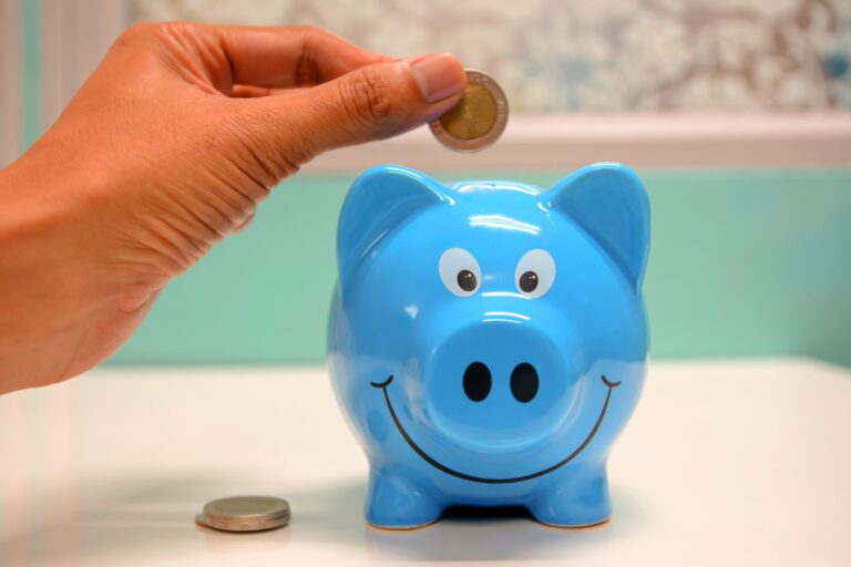 A hand is placing a coin into a blue piggy bank with a smiling face.