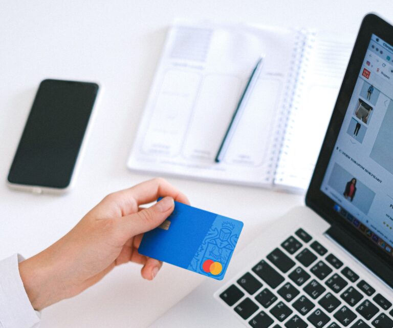 Person holding a blue credit card in their right hand in front of a laptop, with a smartphone, notebook, and pen on a white desk.