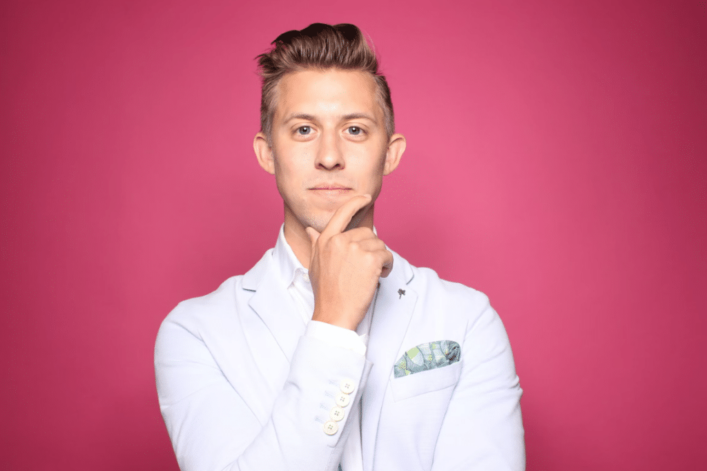 Man in a light suit jacket with a pocket square, standing against a pink background