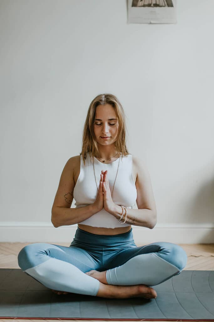 A person sits cross-legged on a yoga mat, wearing a white top and blue leggings, with hands pressed together in a prayer position.