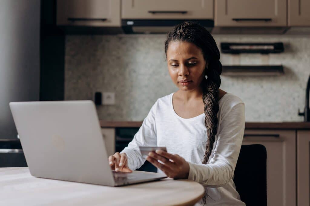 A person with braided hair sits at a kitchen table, using a silver laptop