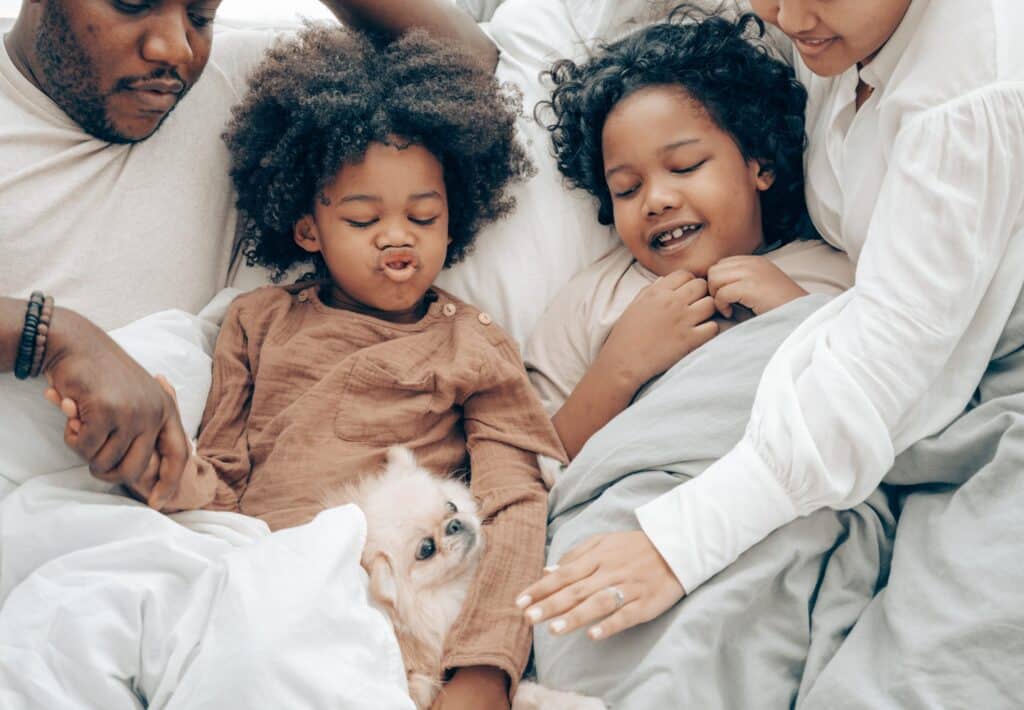 A family of four, including two children, lies on a bed surrounded by white and gray bedding.