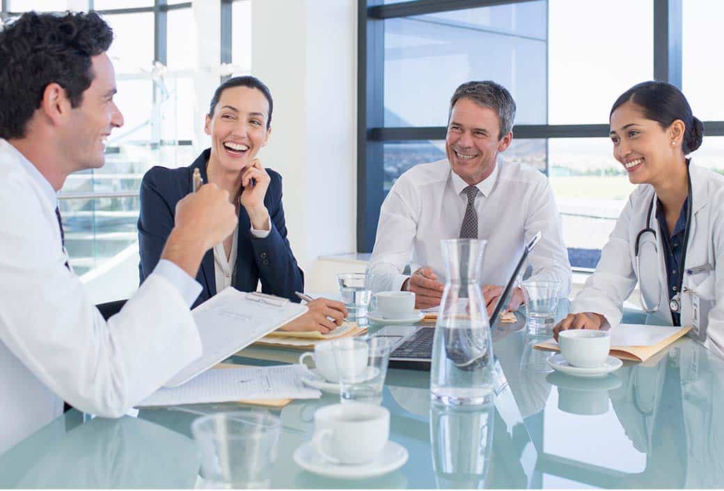 Four professionals, including two in medical attire, sit around a glass table with documents and coffee.