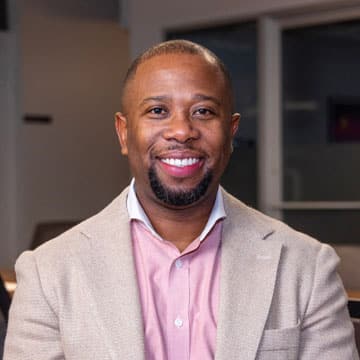 A man wearing a beige blazer and pink shirt smiles while standing indoors with a blurred background.