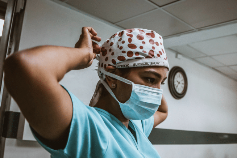 A healthcare worker in blue scrubs and a face mask adjusts her surgical cap with red polka dots in a hospital room.