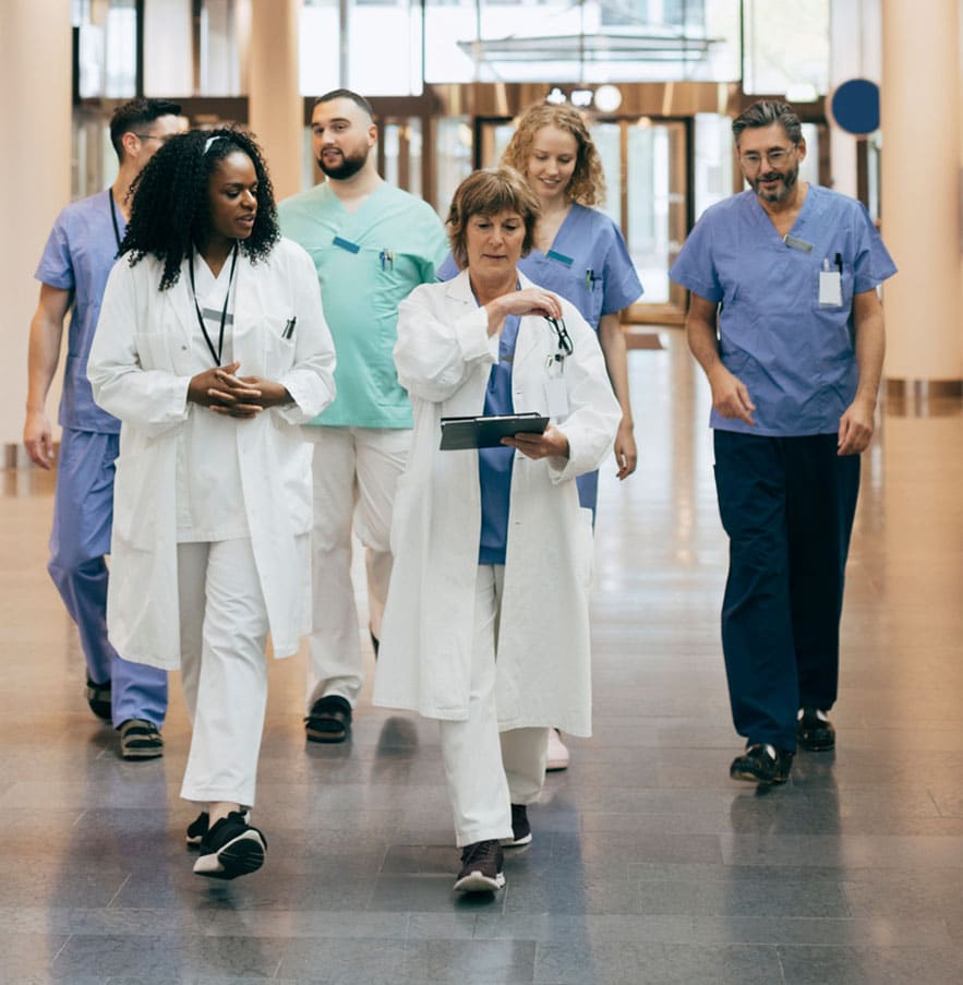 A group of healthcare professionals in scrubs and lab coats walk together in a hospital corridor.