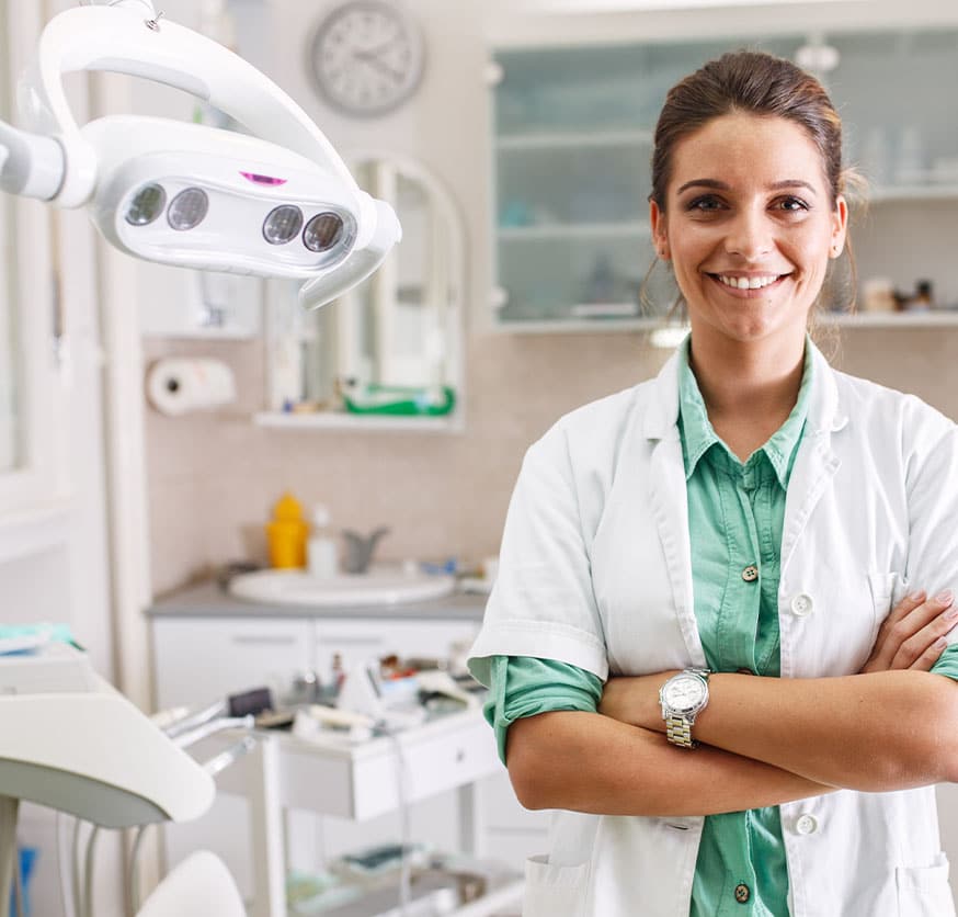 A dentist in a dental clinic, wearing a white coat and green shirt, stands with arms crossed and smiling.
