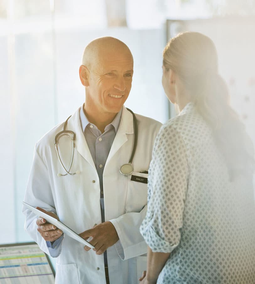 A doctor in a white coat holding a clipboard and smiling at a patient during a consultation in a brightly lit room.