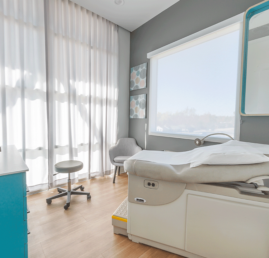 A brightly lit medical examination room with a stool, a gray chair, a teal cabinet, and an exam table.