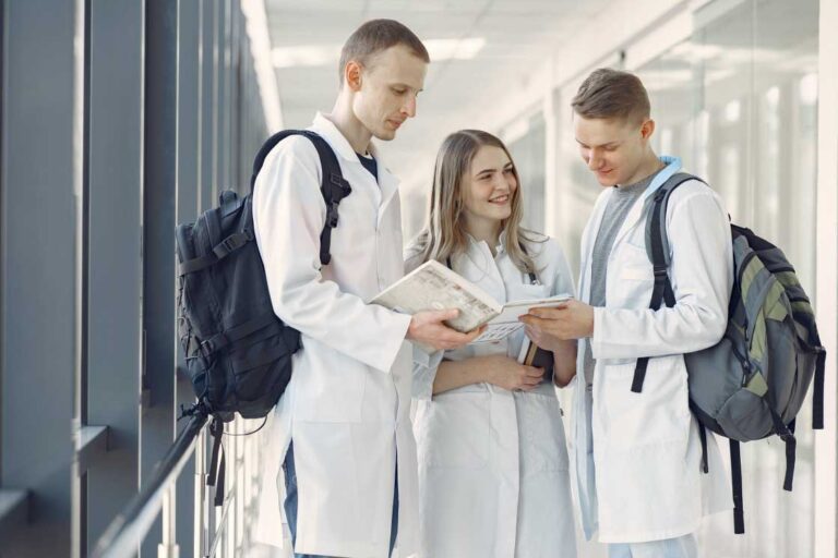 Three students wearing white lab coats and backpacks stand together in a well-lit hallway, looking at books and engaging in conversation.
